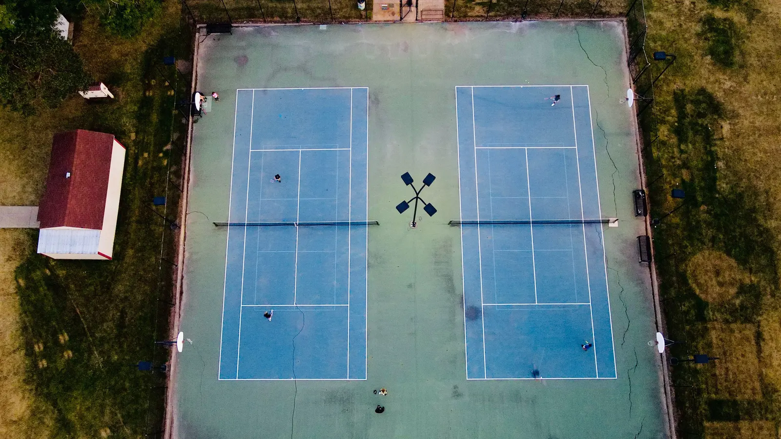 Havadan çekilmiş yan yana iki açık tenis kortu; kortlar ortak bir çevre payını ve ortadaki aydınlatma direğini paylaşıyor.